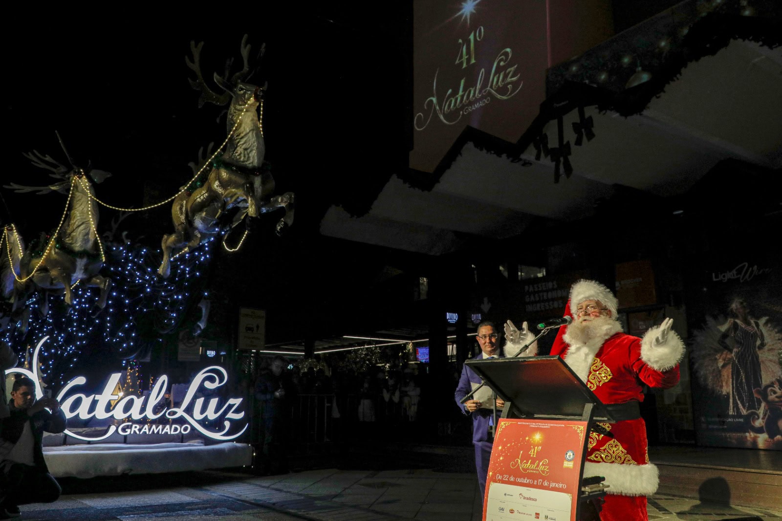 Papai Noel discursa no púlpito ao lado do letreiro iluminado do Natal Luz e do trenó com renas, em Gramado