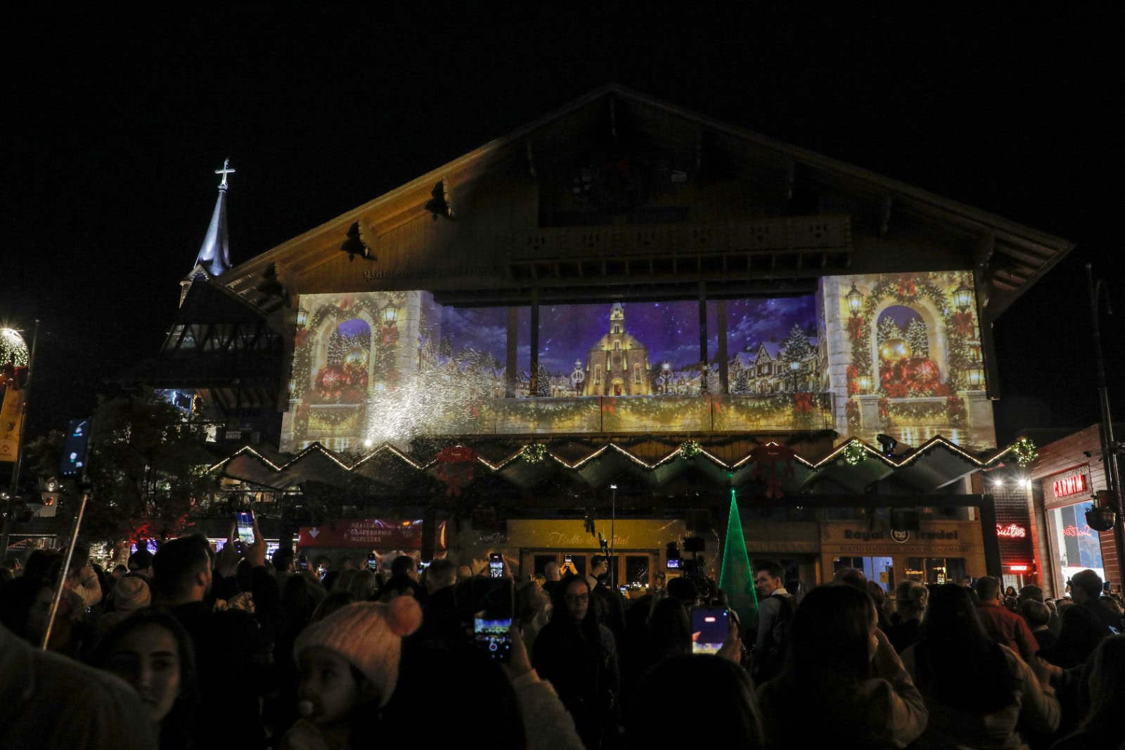 Projeção mapeada de vila natalina na fachada do Palácio dos Festivais, com fogos e público assistindo na rua, em Gramado