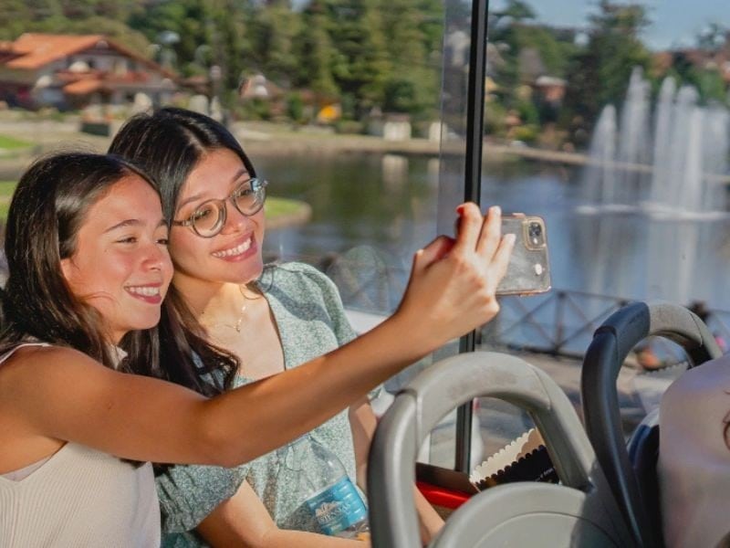 Amigas tirando selfie no Bustour com lago e chafariz ao fundo