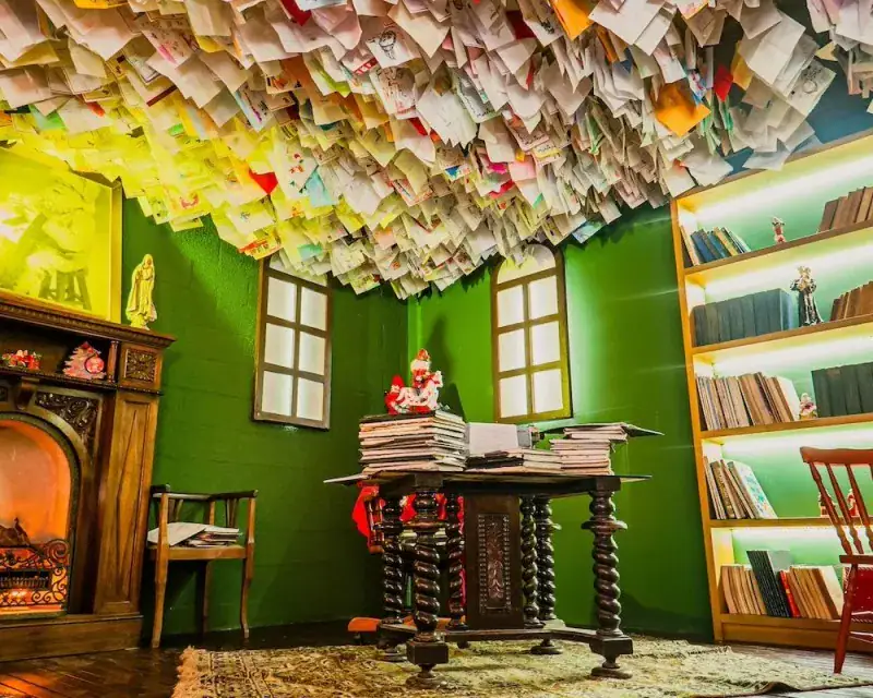 Sala com teto coberto de cartas, lareira e escrivaninha na Aldeia do Papai Noel, em Gramado