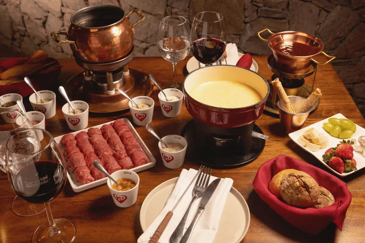 Mesa com fondue de queijo, carne para fondue, panelas de cobre, pães, frutas e taças de vinho no Chalezinho, em Gramado
