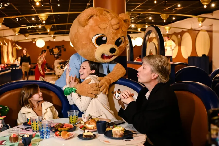 Mascote urso do Clubinho Criamigos abraça família à mesa do café da manhã no Hotel Criamigos, em Gramado