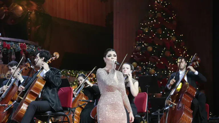 Cantora solista em vestido brilhante com violoncelistas e árvore de Natal iluminada ao fundo, em concerto da Orquestra Sinfônica de Gramado