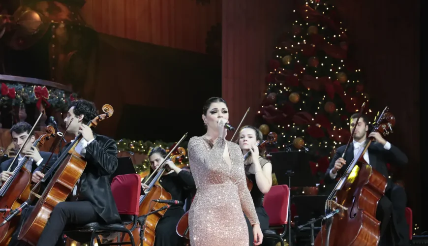 Cantora solista em vestido brilhante com violoncelistas e árvore de Natal iluminada ao fundo, em concerto da Orquestra Sinfônica de Gramado