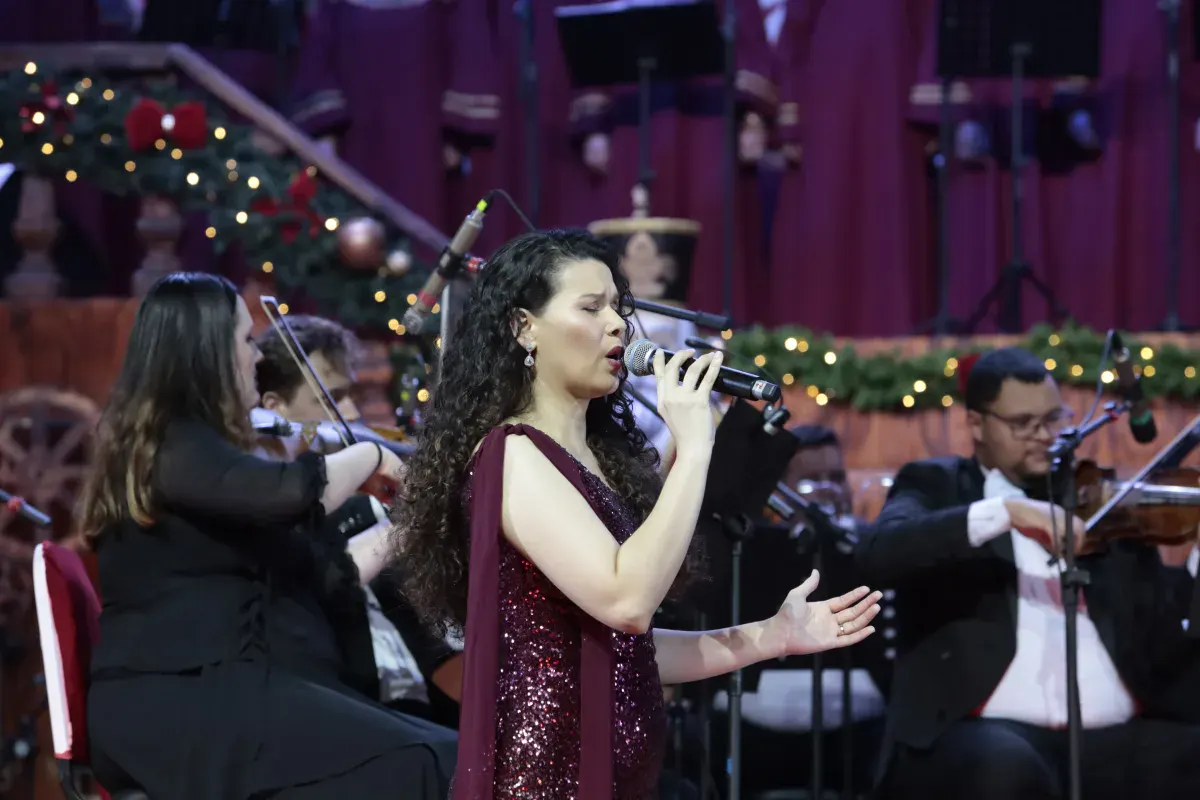Cantora de vestido vinho com brilho cantando ao microfone à frente dos violinistas, em concerto natalino da Orquestra Sinfônica de Gramado